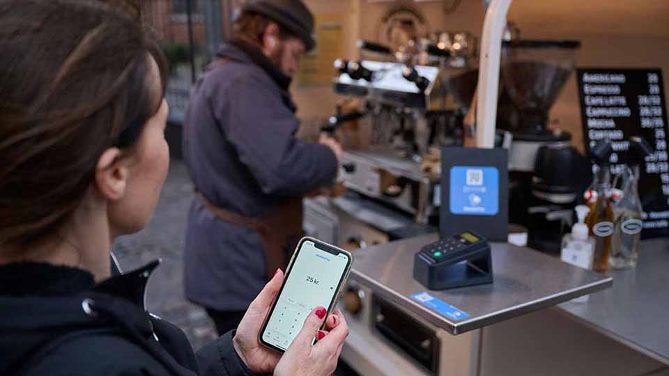 Kvinde står ved en kaffevogn og forbereder mobilbetaling på 25 kr., mens en barista arbejder ved espressomaskinen i baggrunden.