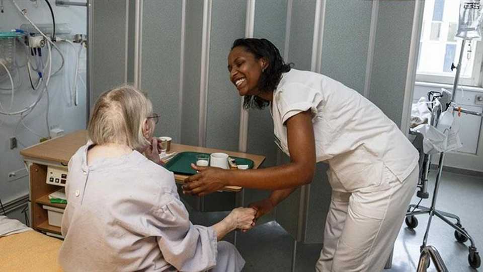 Sygeplejerske smiler og holder en ældre patients hånd på en hospitalsstue med medicinsk udstyr i baggrunden.