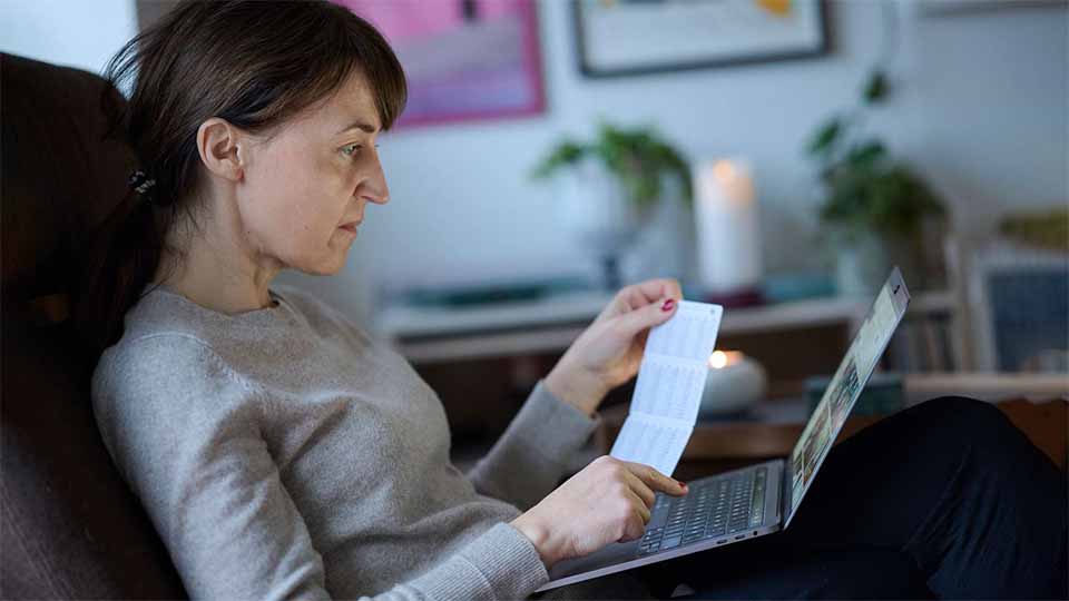 Kvinde sidder i en stol og kigger koncentreret på en bærbar computer, mens hun holder et papir i hånden.
