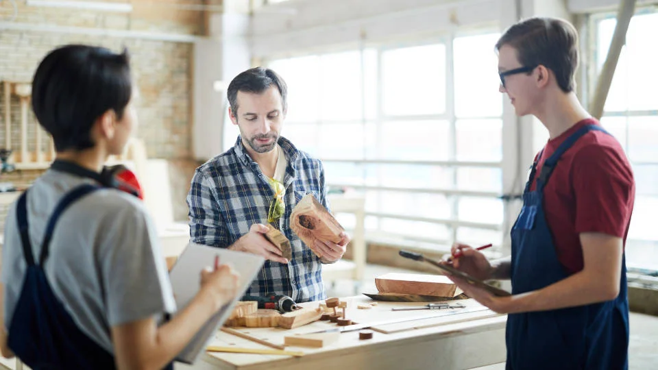 Three people in a woodworking workshop with one person measuring a wooden piece, another taking notes, and a third observing.