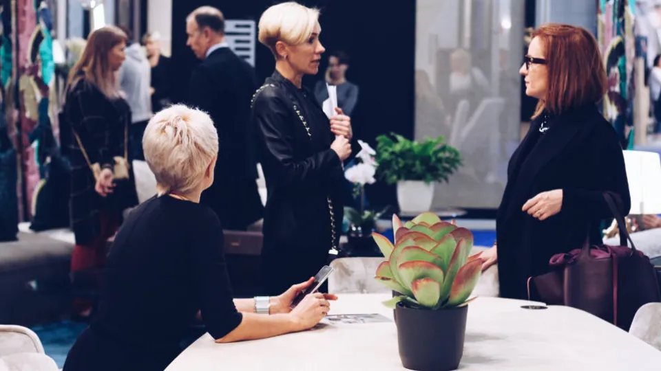 Three women engaging in conversation at a fashion event with a potted plant in the foreground.
