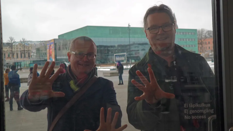 Två leende män som trycker sina händer mot ett fönster med reflektion av en stad i bakgrunden och texten 'Ej genomgång No access' synlig.