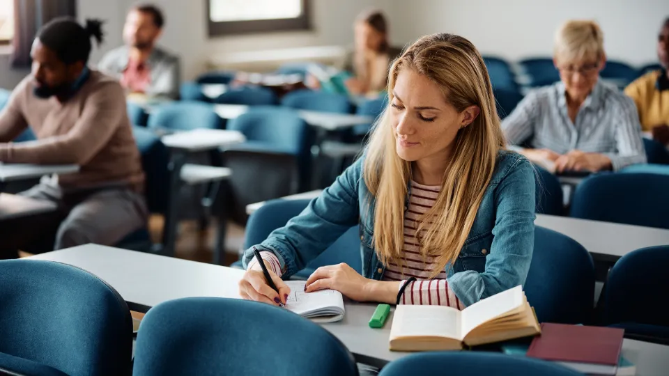 Kvinna skriver anteckningar i ett klassrum med andra studenter i bakgrunden.
