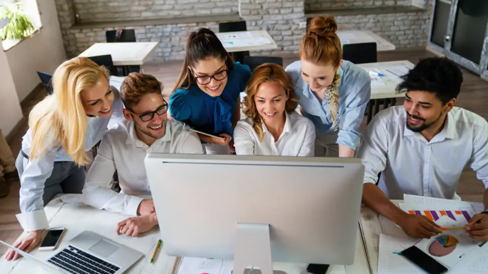 A diverse group of six colleagues happily collaborating around a computer in a modern office.