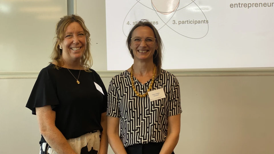 To kvinder smilende foran en præsentationsskærm under en workshop eller konference.