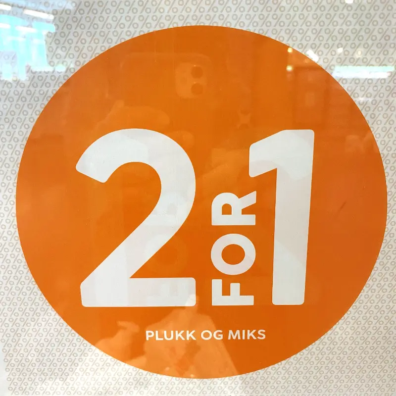Orange cirkulært skilt med teksten "2 for 1 plukk og miks" på en butiksdør.