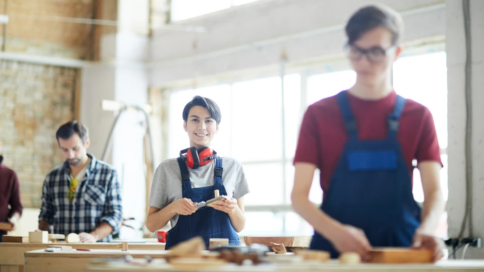 Unge mennesker i arbejdsdragter arbejder i et træværksted, med fokus på en smilende person i forgrunden med hovedtelefoner om halsen.
