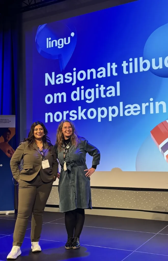 To kvinner står foran en storskjerm på en konferanse. På skjermen står teksten "Lingu - Nasjonalt tilbud om digital norskopplæring". De smiler og har konferansemerker rundt halsen.