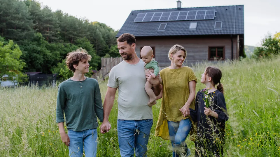 "Familj i naturskön miljö framför ett hus med solpaneler på taket, omgiven av en grön äng och omgärdad av skog. De håller varandra i händerna och ser glada och harmoniska ut."