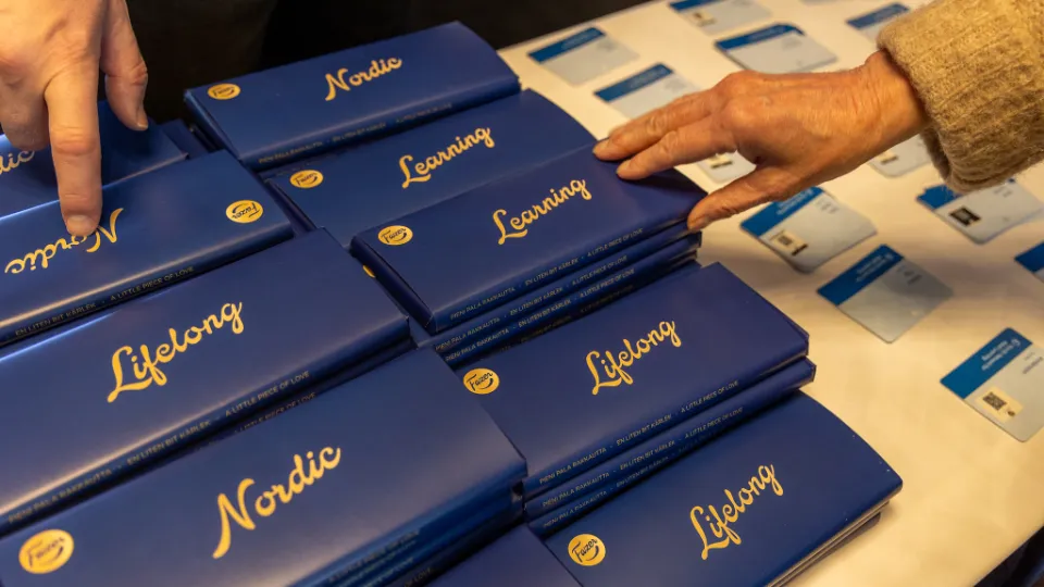 Dette billede viser en række blå chokoladebarer, pænt stablet på et bord. Hver bar har gylden skrift med ordene "Nordic," "Learning" og "Lifelong," hvilket sandsynligvis henviser til et arrangement eller koncept om livslang læring i Norden. Man kan også se hænder, som tager eller peger på chokoladebarerne, hvilket antyder, at de bliver delt ud ved en event. I baggrunden ligger der kort eller navneskilte, som kan indikere en konference eller et seminar. Den blå og gyldne farve giver et elegant og professionelt udtryk.