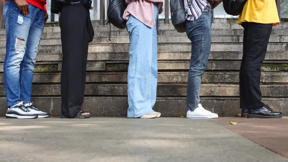 Billedet viser en gruppe personer stående i kø. Fokus er på deres ben og fødder, som er klædt i forskelligt tøj og fodtøj. Trinene i baggrunden antyder en offentlig eller udendørs placering, og personerne har rygsække, hvilket kan tyde på en hverdagssituation som venten på transport eller adgang til en bygning.