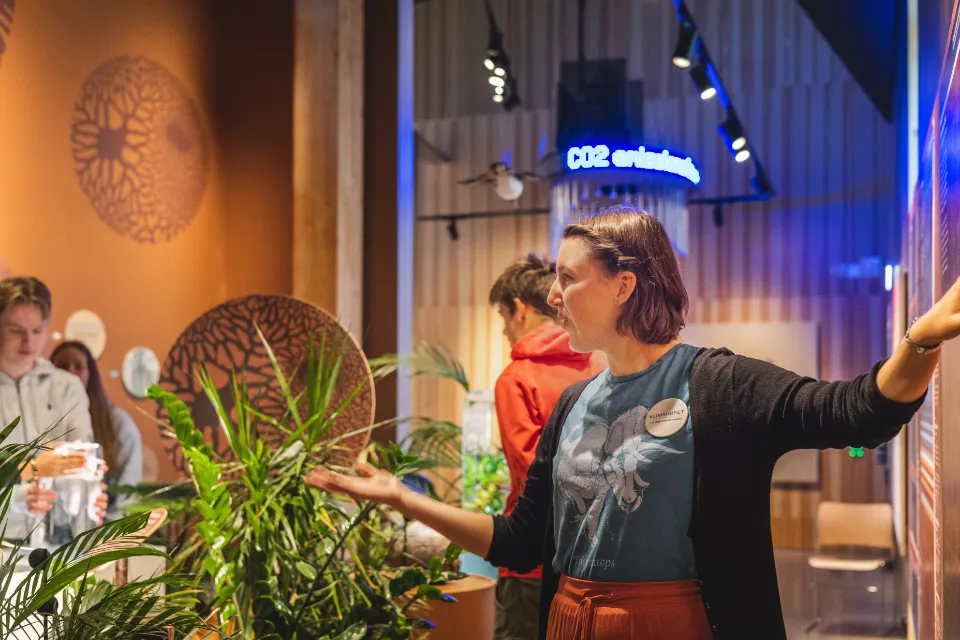En kvinna som presenterar information i en växtfylld utställningsmiljö med fokus på hållbarhet.