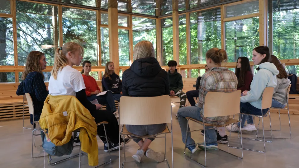 En grupp personer som sitter i en cirkel i ett ljust rum omgivet av fönster och grönska.