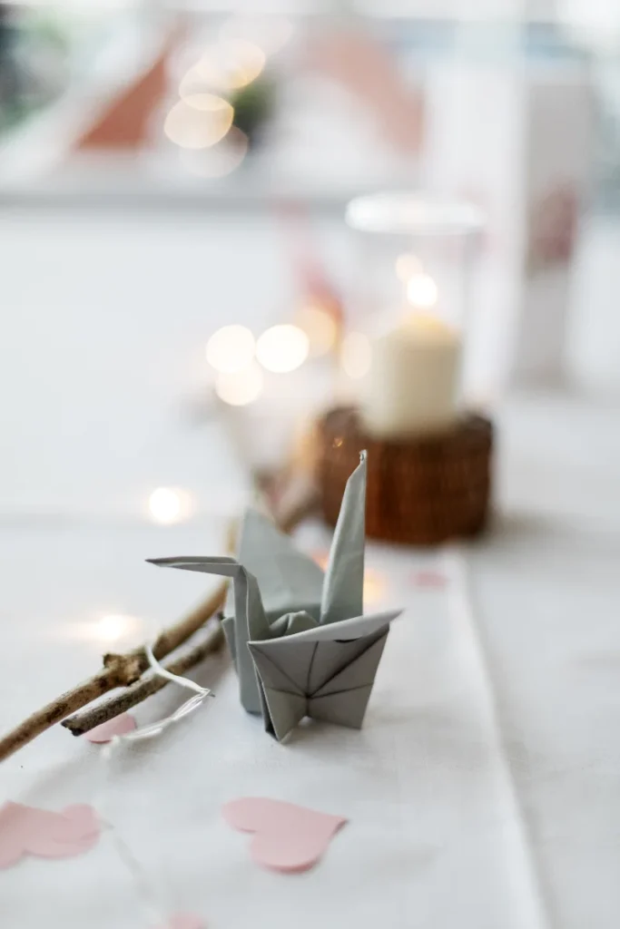 Närbild av en origami-trana på ett dukat bord med ljus, rosa hjärtformade dekorationer och en tänd ljuslykta i bakgrunden.