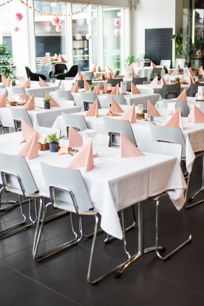 Ljusa dukade bord med vikta servetter i rosa och dekorationer i en ljus, modern lokal med stora fönster.