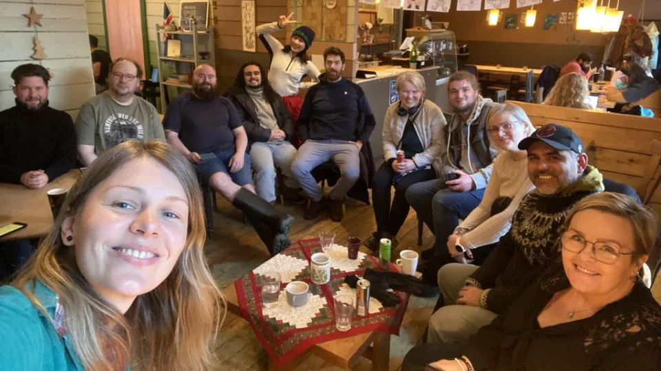 A group of people gathered around a table in a cozy café, smiling for a group photo. The café has warm wooden decor and festive holiday accents.