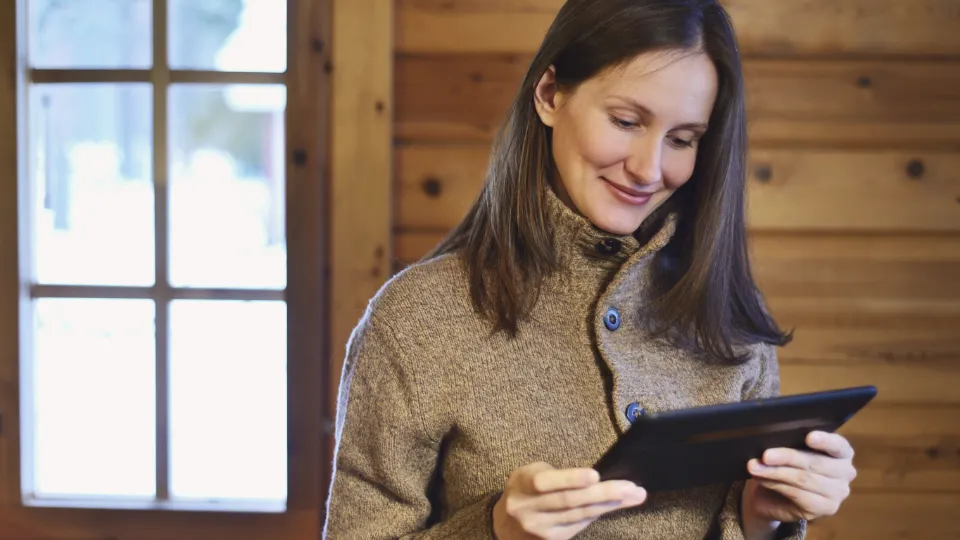 A smiling woman in a cozy wool sweater uses a tablet inside a wooden cabin near a window.