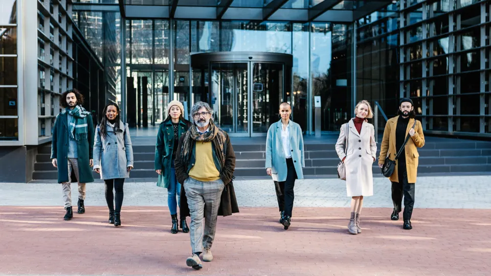 En gruppe mangfoldige forretningsfolk iført stilige vinterklær går selvsikkert ut av en moderne kontorbygning med glassfasade.