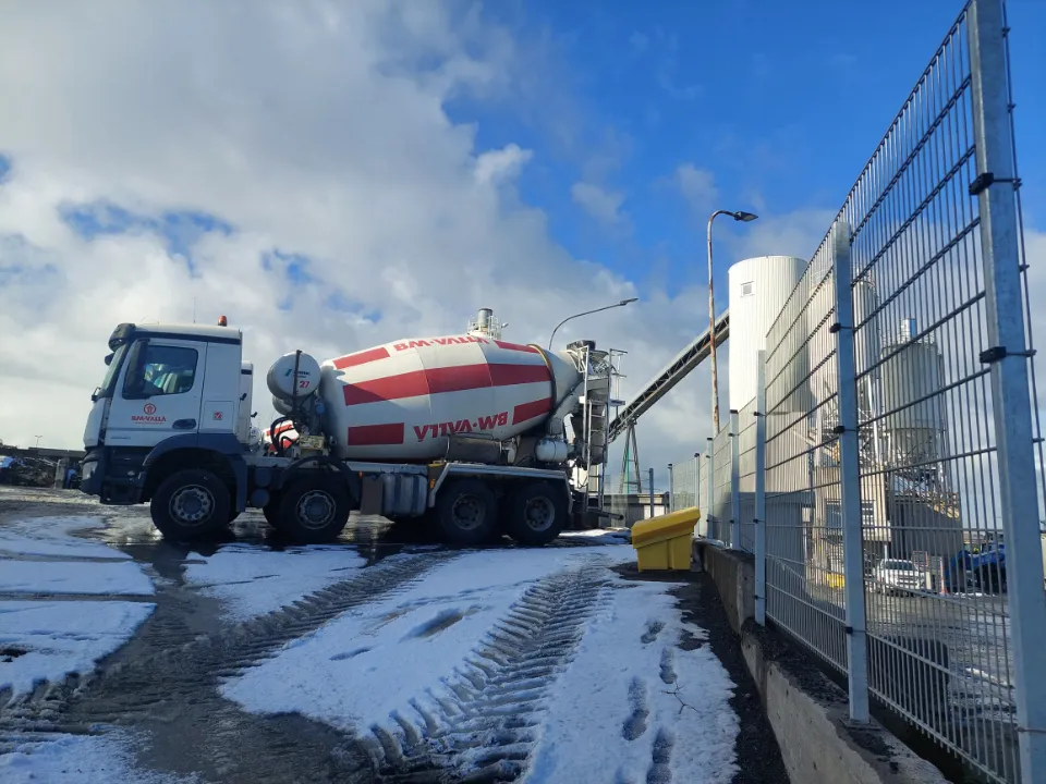 En betongbil med røde og hvite striper fra BM-Valla står parkert ved en fabrikk med snø og is på bakken.