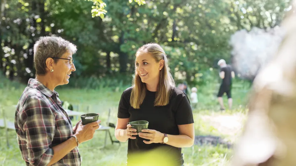 To kvinder står i en skovlysning og snakker, mens de holder kopper. Solen skinner gennem træerne, og i baggrunden ses flere personer, herunder et barn og en mand i sort tøj.