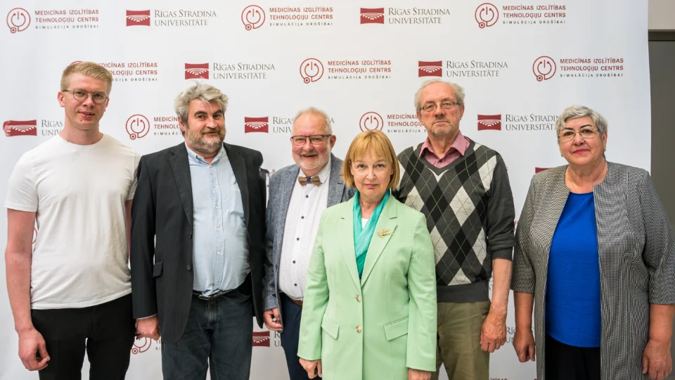 A group of six people posing in front of a backdrop with "Rīga Stradiņš University" and "Medical Education Technology Center" logos. They are dressed in a mix of casual and formal attire, smiling at the camera.