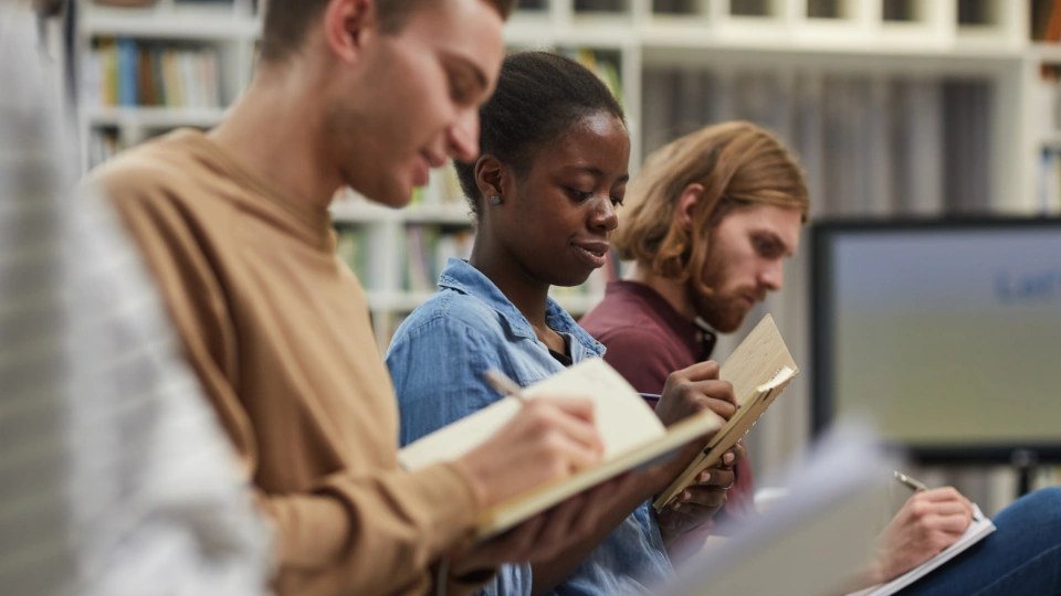 Tre unga vuxna sitter i ett bibliotek och skriver i anteckningsböcker med fokus.