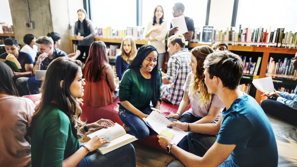 Studenter som sitter i grupp och studerar tillsammans i ett bibliotek, omgivna av böcker och naturligt ljus.