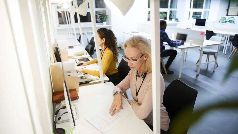Kvinder og mænd arbejder ved skriveborde i et moderne kontormiljø med computere og dokumenter.