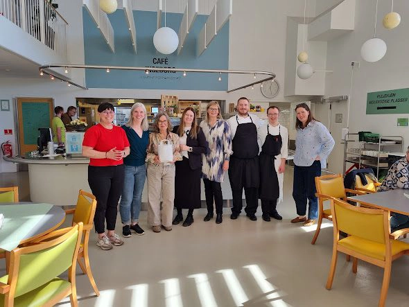 Gruppe mennesker samlet i et lyst kafélokale med moderne innredning, smilende foran disken i et sosialt miljø.