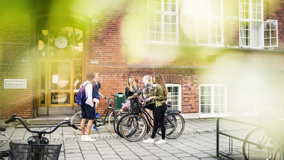 En gruppe unge står med sykler utenfor en rød murbygning med store vinduer og en inngangsdør med klokke over.