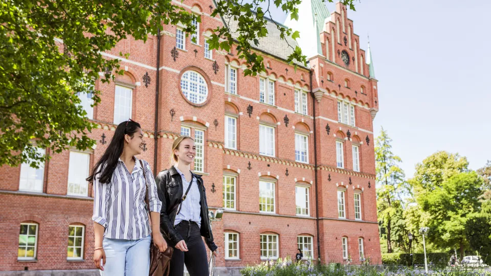Två unga kvinnor promenerar framför en historisk röd tegelbyggnad med gröna tak i en lummig parkmiljö.
