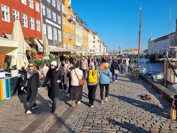 Mange mennesker går langs havnepromenaden i Nyhavn, København, omgitt av fargerike bygninger og båter.
