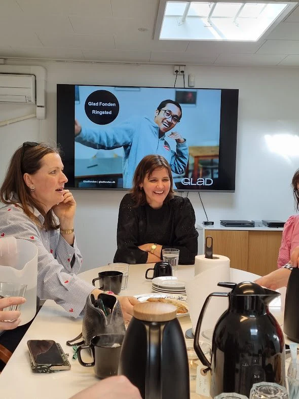 Kvinner sitter rundt et bord i et møterom med kaffekanner og skjerm i bakgrunnen som viser "Glad Fonden Ringsted".