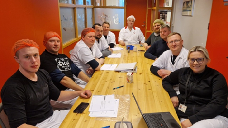 Gruppe med fabrikkarbeidere og ansatte i arbeidsklær og hårnett samlet rundt et møtebord med dokumenter og en bærbar PC.