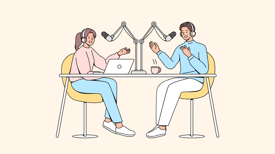 Two people recording a podcast at a table with microphones, headphones, a laptop, and a coffee cup.