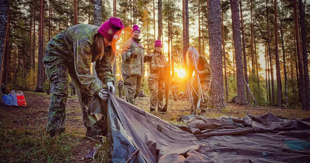 Grupp i militärkläder och lila mössor sätter upp tält i en skog vid solnedgång.