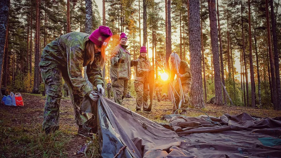 Grupp i militärkläder och lila mössor sätter upp tält i en skog vid solnedgång.