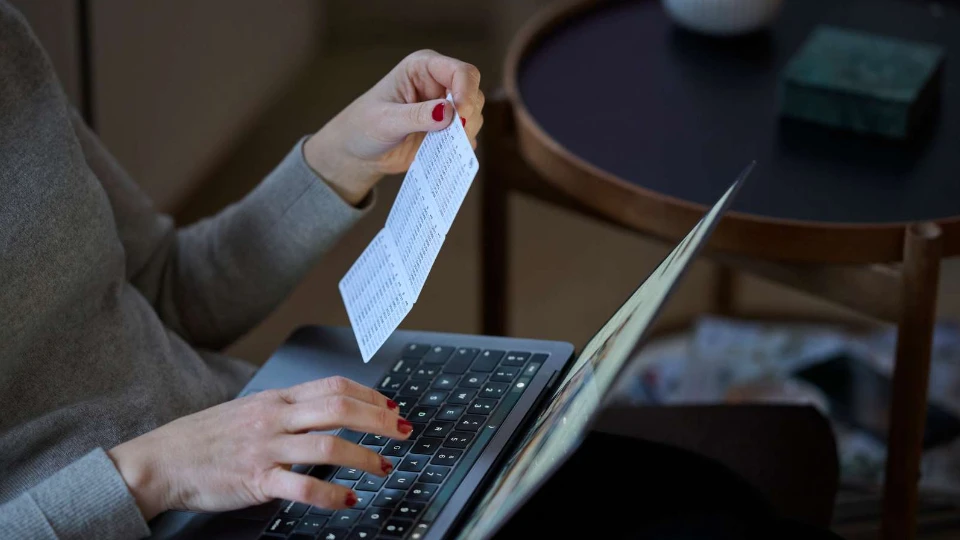 Person typing on a laptop while holding a printed document with codes or numbers, sitting at a table indoors.