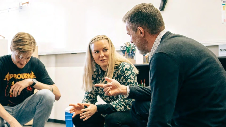 Underviser i dialog med to voksne studerende i et klasseværelse, med tekst om højere kvalitet, fleksibilitet og livslang læring.