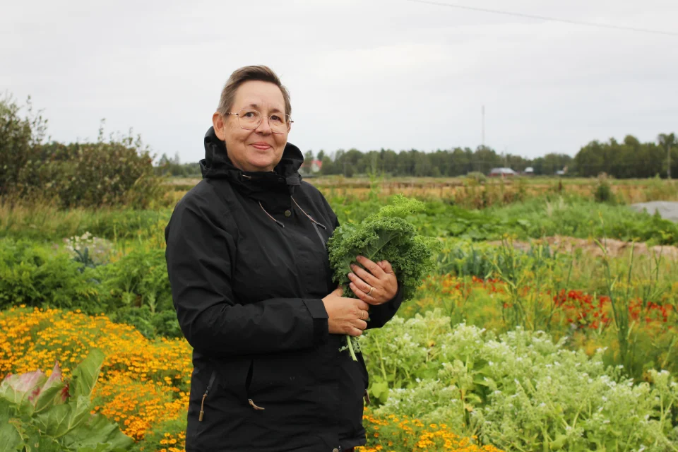 Kvinna i svart jacka håller grönkål i en färgstark trädgård med blommor och grönsaker på landet.