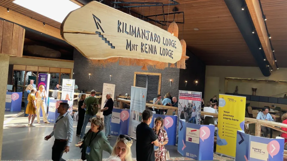 People networking and visiting exhibition booths at an indoor business event held at Kilimanjaro Lodge, with visible banners and informational displays.
