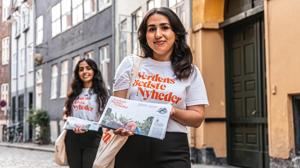 To kvinder iført t-shirts med teksten "Verdens Bedste Nyheder" uddeler aviser på en brostensbelagt gade i en historisk bydel.