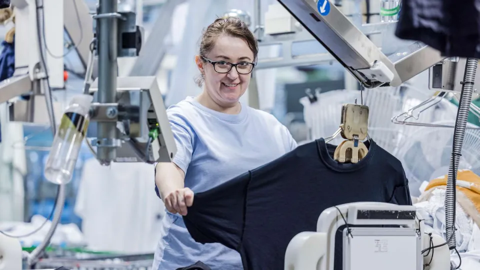 Arbetslivsorienterad yrkesutbildning sätter fart på den gröna omställningen Kvinna i produktionsmiljö håller upp en svart T-shirt framför automatiserad maskinutrustning.