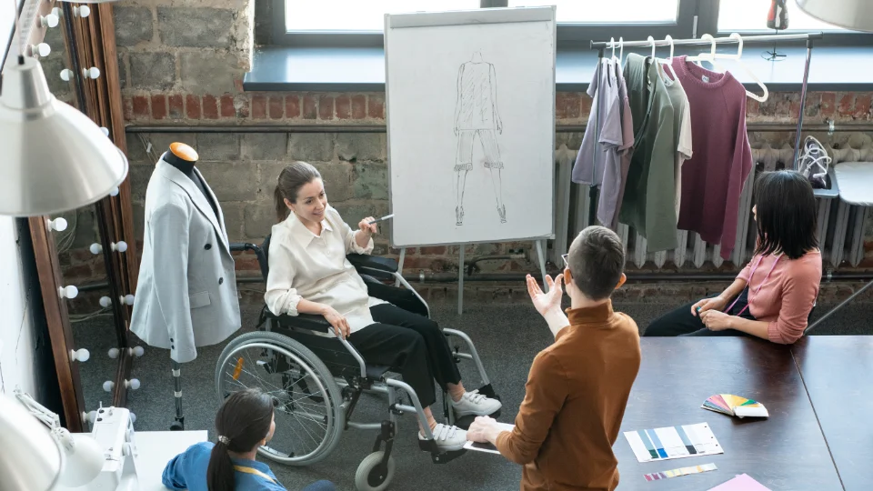 Folkhögskolans yrkeskurser främjar etablering på arbetsmarknaden Kvinna i rullstol håller modepresentation framför team i designstudio med klädhängare, provdocka och skiss på whiteboard.