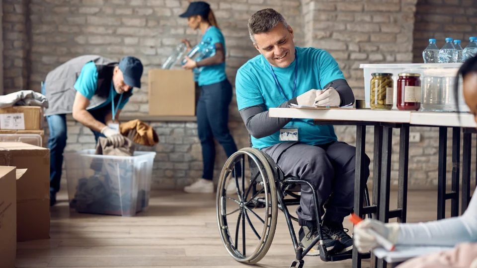 Forsøk med lønnstilskudd for å få flere unge voksne i jobb Frivillige som sorterer donasjoner i et hjelpesenter, inkludert en mann i rullestol som skriver ved et bord.