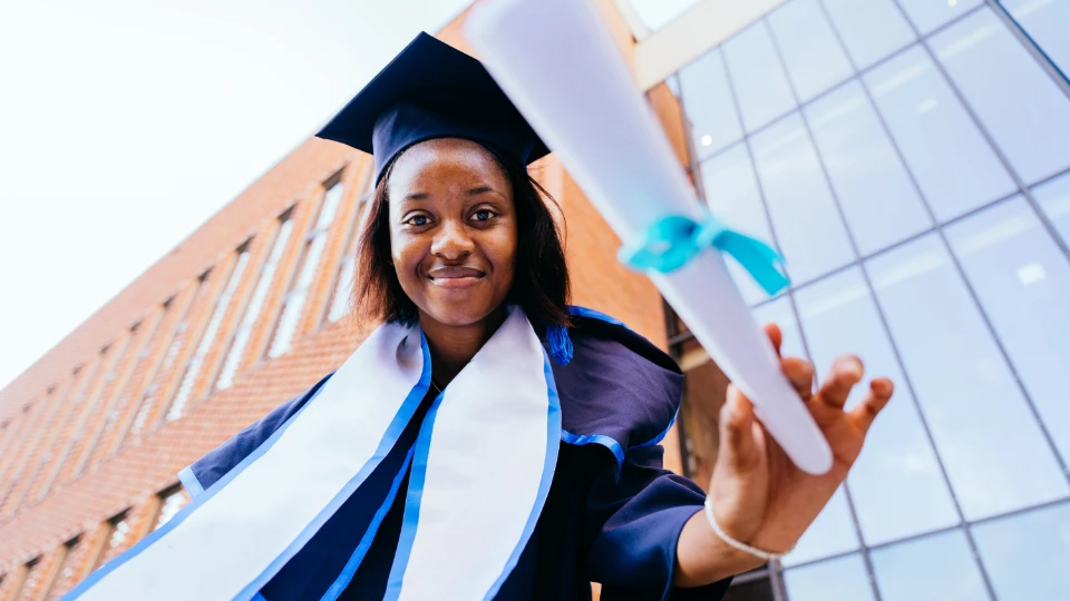 Ung kvinna i examensdräkt håller ett diplom framför en modern skolbyggnad.