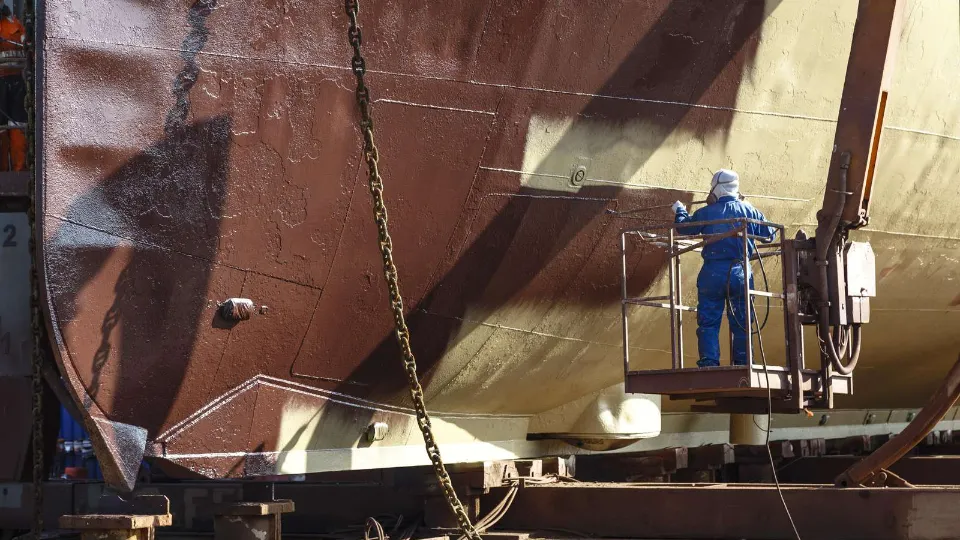 Person målar ett fartygsskrov med skyddsutrustning på ett varv.