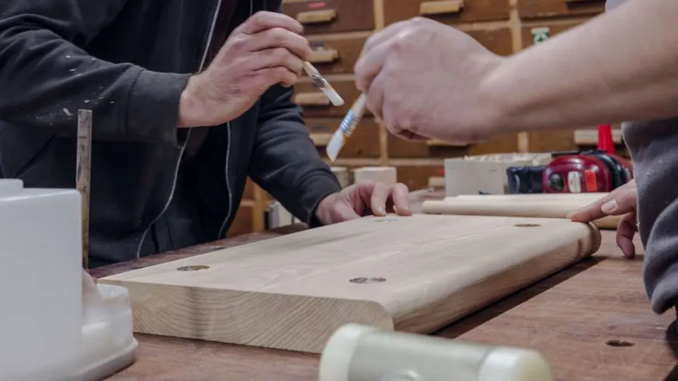 Hvorfor unge fravælger erhvervsuddannelser To personer arbejder med træbearbejdning og påfører lim på et stykke lyst træ i et værksted.