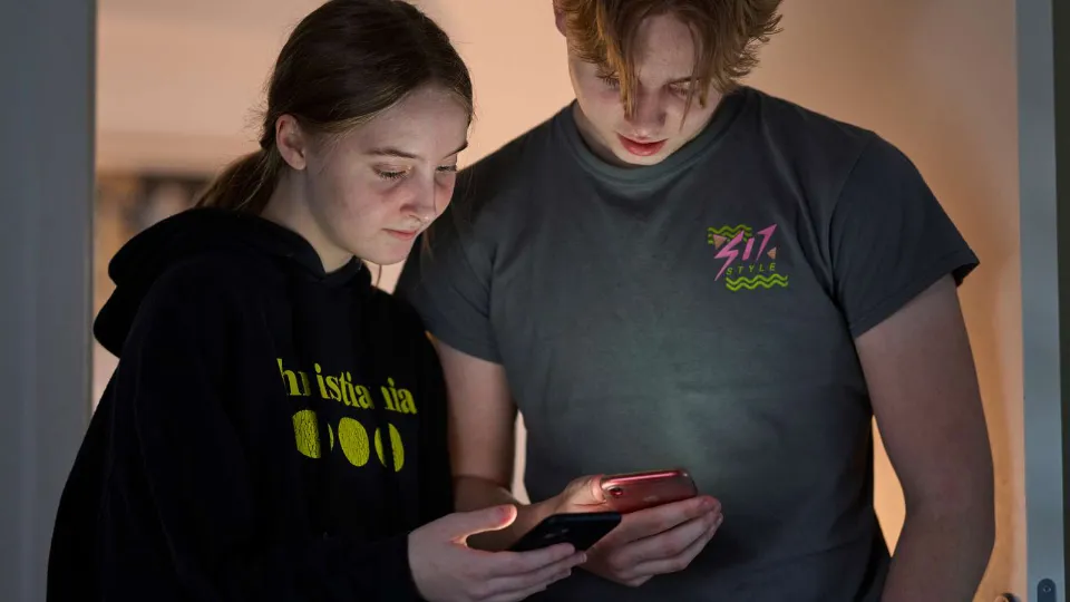 To ungdommer står sammen og ser på en mobiltelefon i dempet belysning.
