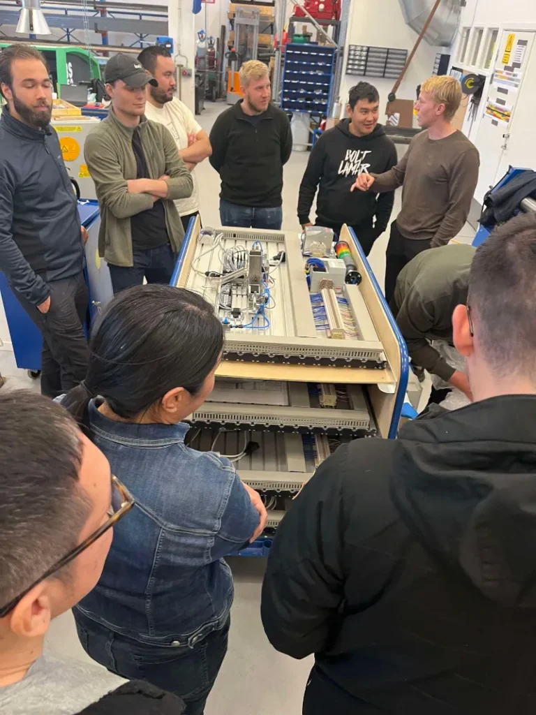 En gruppe mennesker samlet rundt et teknisk kontrollpanel i et verkstedmiljø, hvor én person forklarer komponentene.